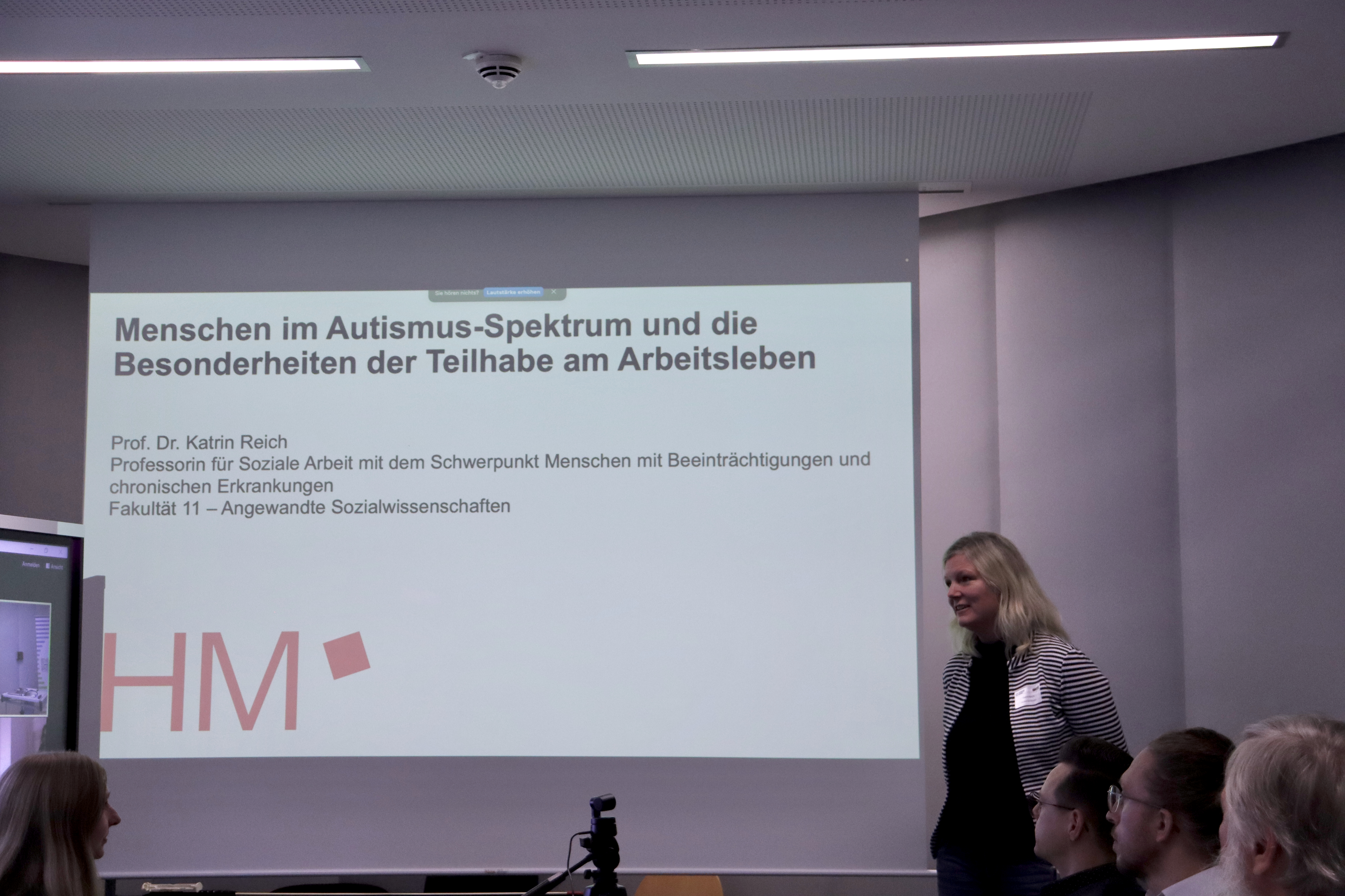 Photo of a PowerPoint presentation on a screen entitled "People on the autism spectrum and the special features of participation in working life". To the right of the presentation is a female person (Katrin Reich) speaking.