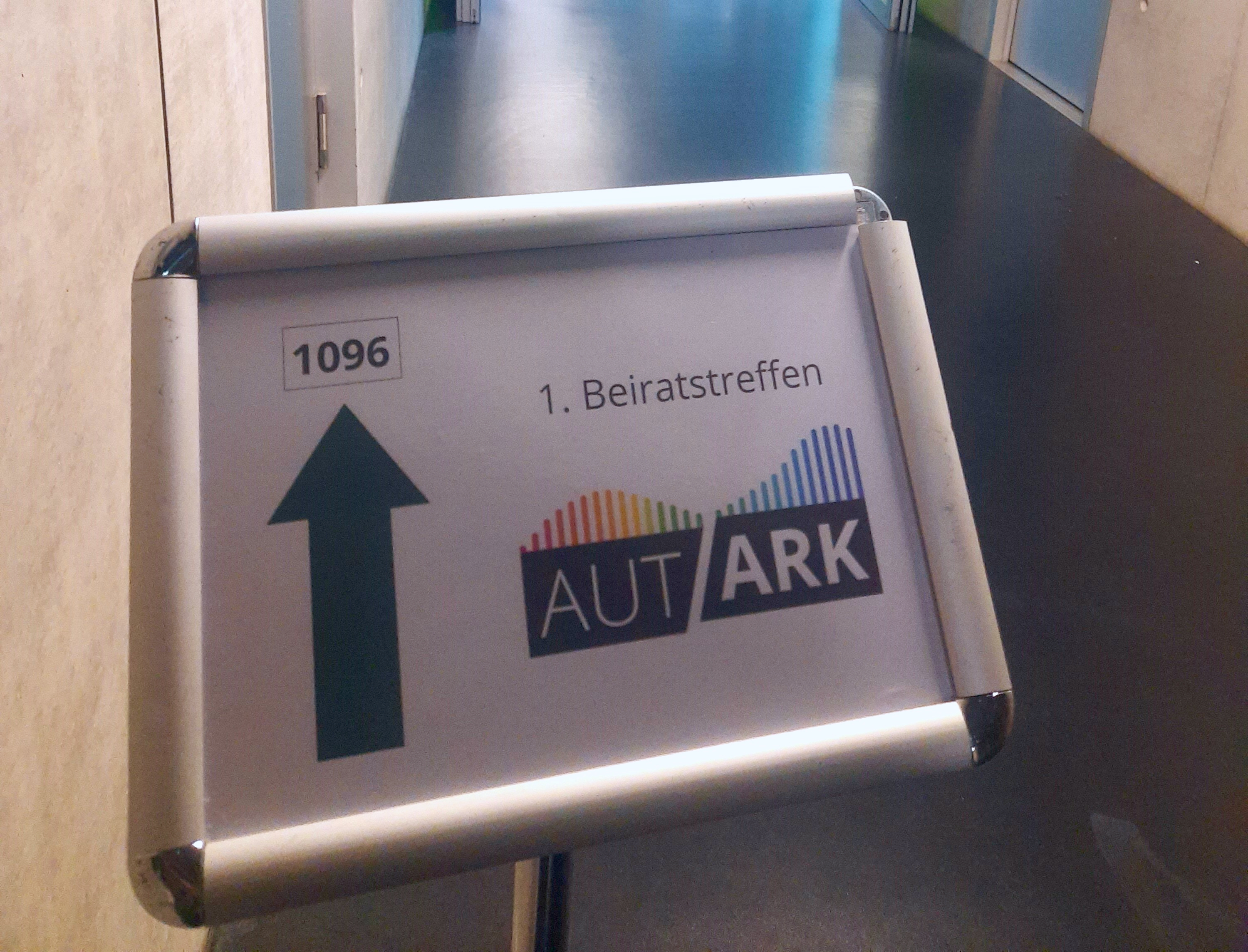 Aufstellschild in einem Flur mit einem Pfeil nach vorn und der Beschriftung "1096" Rechts daneben das Logo von Autark und "1. Beiratstreffen"