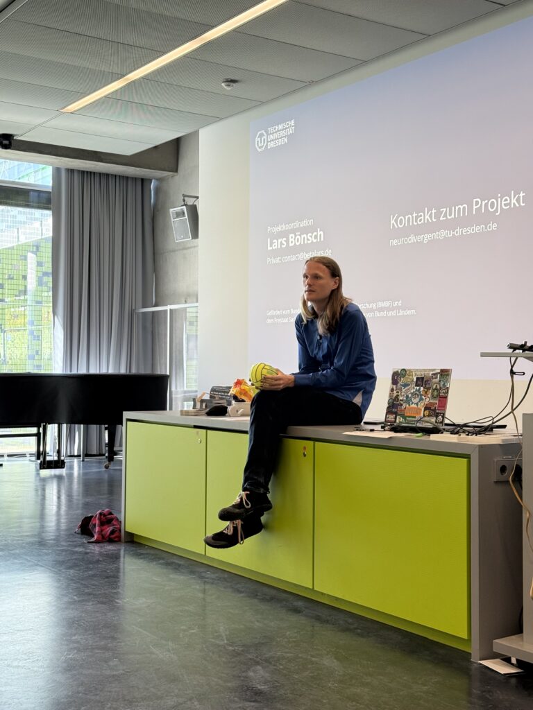 Lars Bönsch sitzt auf dem langen Pult vorn im Raum und spricht. Folien an der Wand im Hintergrund mit seinem Namen und Kontakt